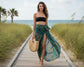 Woman in a black top and patterned sarong standing on a wooden path with grass and ocean in the background