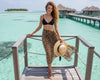 Woman in a leopard print sarong and black bikini top standing on a wooden dock with turquoise water and huts in the background.
