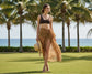Woman in a leopard print sarong and black top standing on grass with palm trees and ocean in the background