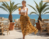 Woman in a white sarong top and tiger print sarong holding a drink on a patio with palm trees and ocean view.