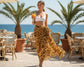 Woman in a white sarong top and tiger print sarong holding a drink on a patio with palm trees and ocean view.