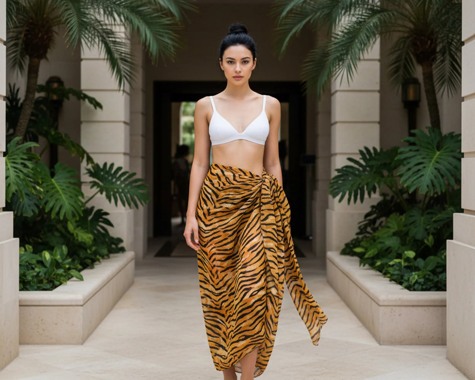 Woman in a white sarong top and tiger striped sarong standing in a tropical setting with plants and stone architecture.