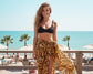 Woman in black bikini top and tiger print sarong standing on a beach with palm trees and ocean in the background.