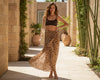 Woman wearing a leopard print sarong and black top in an outdoor setting with plants and stone walls.