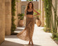 Woman wearing a leopard print sarong and black top in an outdoor setting with plants and stone walls.