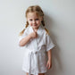 Toddler girl in a white muslin robe with braided hair, smiling and posing against a light wall