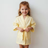 Little girl wearing a soft yellow muslin robe with a tie belt, smiling while standing indoors.