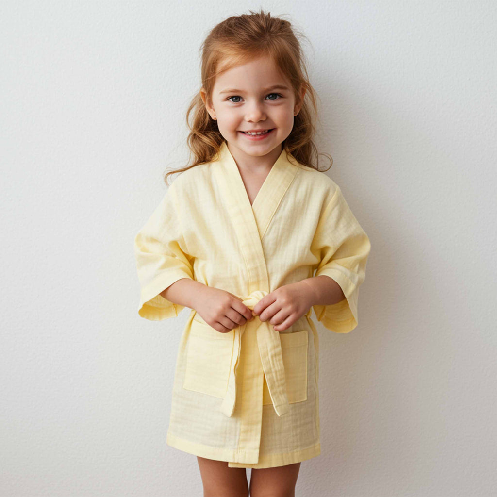 Little girl wearing a soft yellow muslin robe with a tie belt, smiling while standing indoors.