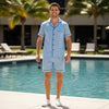 Man in light blue shirt and shorts standing by a pool with palm trees in the background