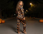 Woman wearing a Halloween-themed onesie with pumpkin pattern in a dark outdoor setting with pumpkins.
